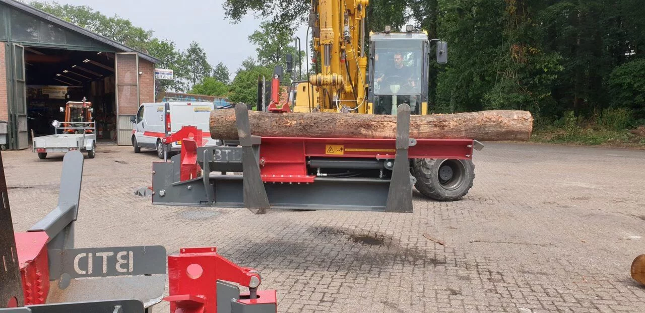 Hydraulische houtklover van BTD tilt dikke boomstam op bedrijventerrein met graafmachine, klaar voor houtverwerking.
