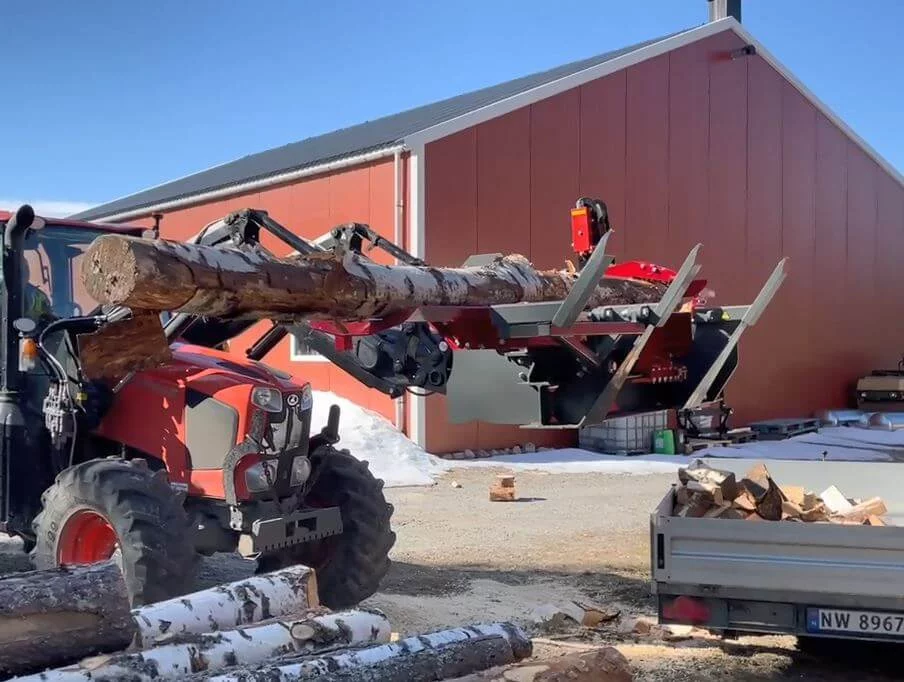 Rode tractor met grijparm laadt een dikke boomstam in een BTD zaagkloofmachine bij een houtverwerkingslocatie, met aanhanger vol brandhout op de voorgrond.