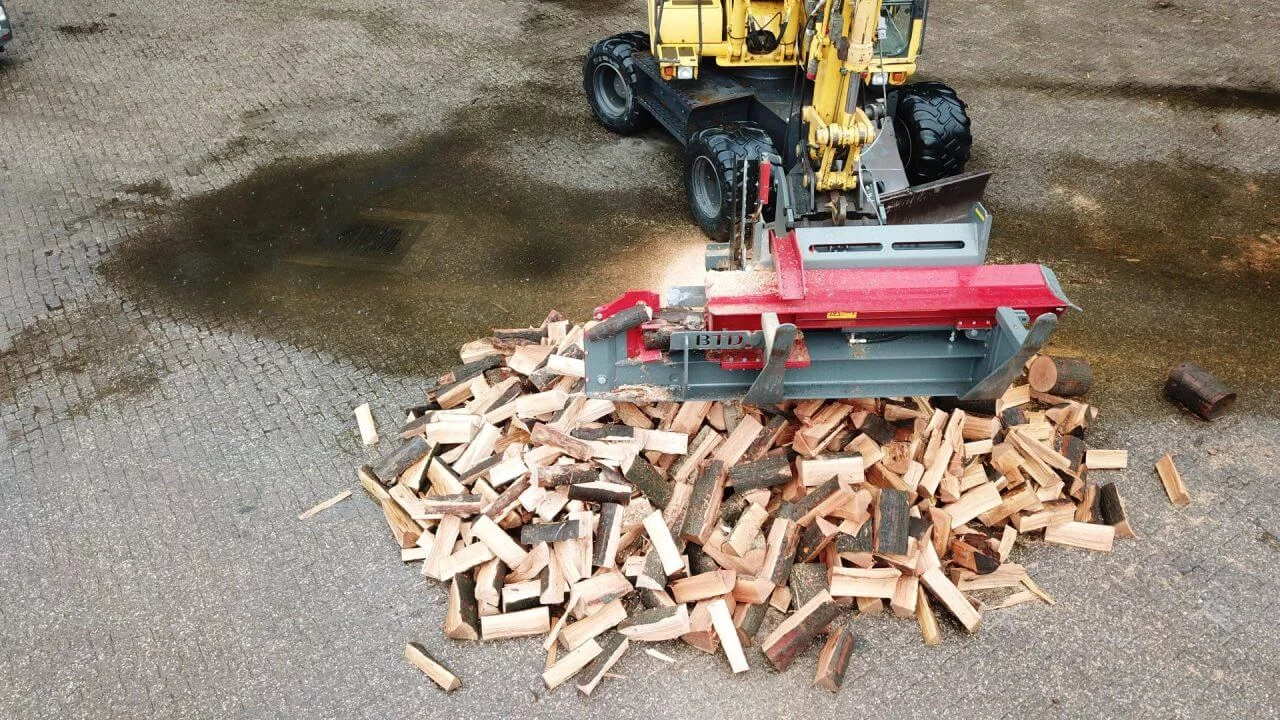 Hydraulische houtklover aan het werk, aangedreven door een gele wiellader, met een hoop vers gekloofd brandhout op een nat bestraat terrein.