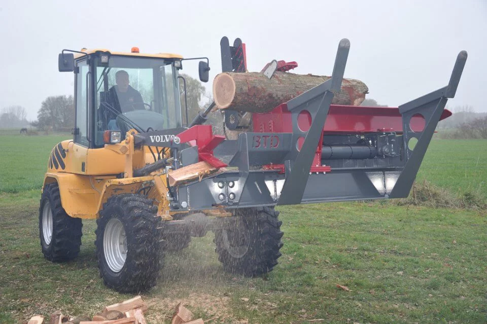 Volvo shovel met gemonteerde BTD houtkloofmachine in actie op een grasveld, waarbij boomstammen worden gekloofd en houtblokken op de grond vallen.