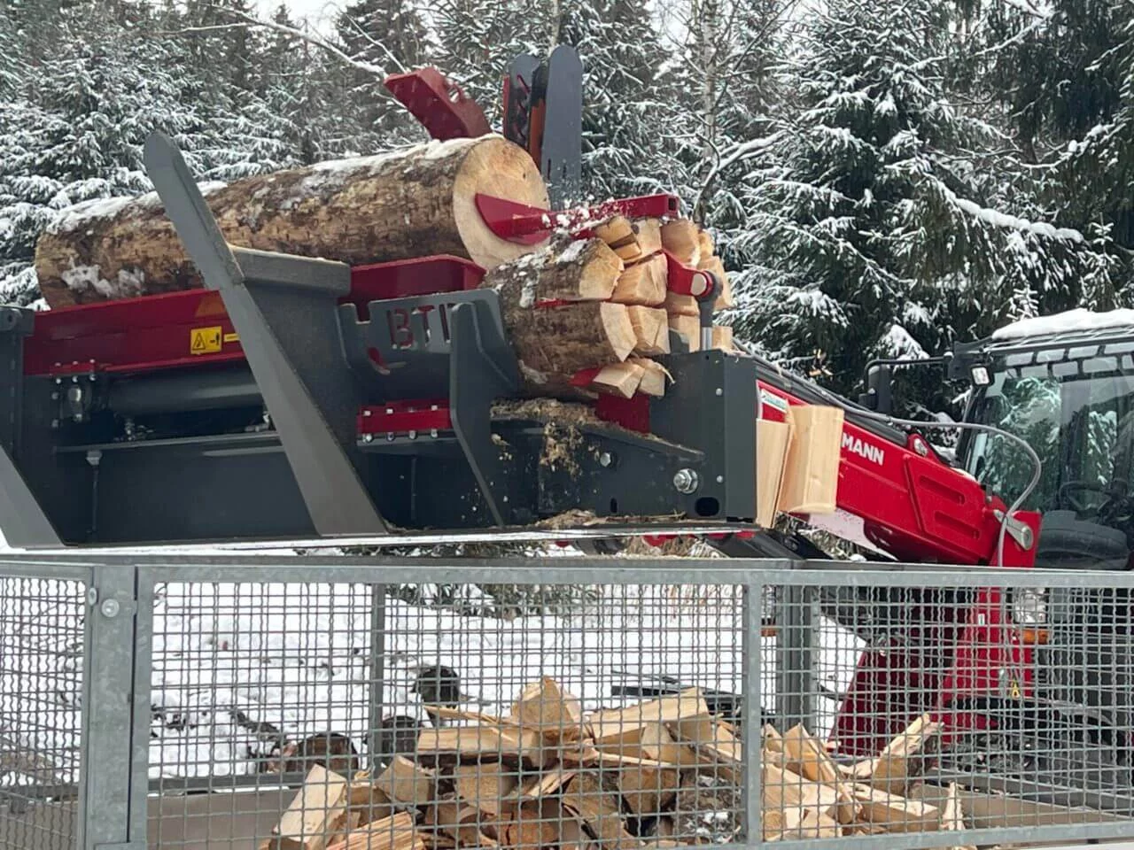 BTD houtkloofmachine verwerkt boomstammen tot brandhout in een besneeuwd bos, met een rode Weidemann shovel op de achtergrond en een metalen opvangbak op de voorgrond.