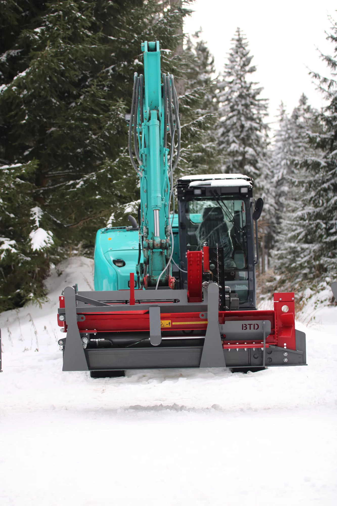 BTD houtklover bevestigd aan een turquoise graafmachine, werkend in een besneeuwd bosgebied.