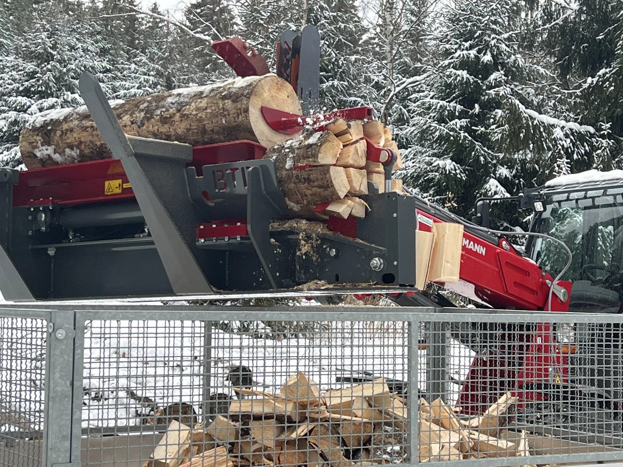 Houtklover van BTD die boomstammen splijt tot brandhout, aangestuurd door een rode loader in een besneeuwd bosgebied.